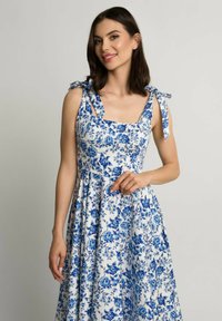 Kwiatowa sukienka na białym tle z niebieskim wzorem kwiatów, z kwadratowym dekoltem, związanymi ramiączkami oraz rozkloszowanym dołem.