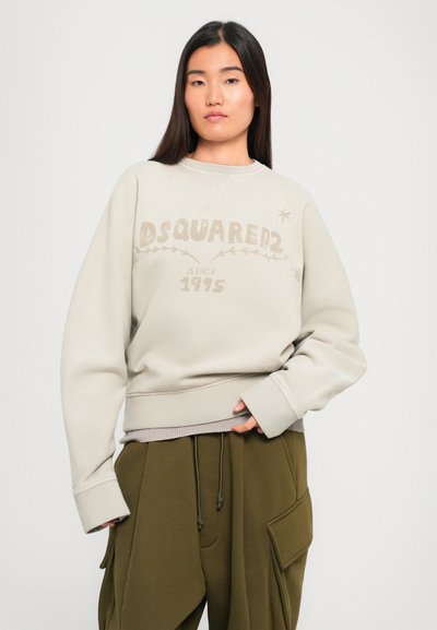 Woman wearing light beige sweatshirt with "DSQUARED2 since 1995" text and olive green cargo pants, standing against a white background.