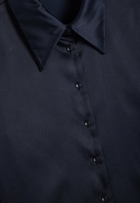 Camisa de color azul marino con cuello puntiagudo y seis botones negros en el frente; textura suave y diseño minimalista.