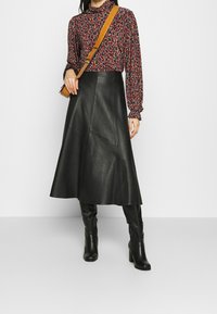 Schwarzer Leder-Midi-Rock mit ausgestellter Silhouette und sichtbaren Nahtdetails. Kombiniert mit einer langärmligen gemusterten Bluse und knielangen Stiefeln.