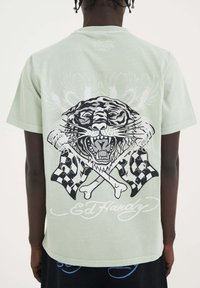 Světle zelené bavlněné tričko s velkým černým grafickým motivem tygra se zkříženými vlajkami a kostmi, značkované "Ed Hardy" pod ním.
