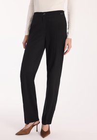 Kobieta ubrana w czarne dopasowane spodnie z zapięciem na guziki, w połączeniu z białą bluzką z długim rękawem oraz brązowymi szpilkami z odkrytym palcem.