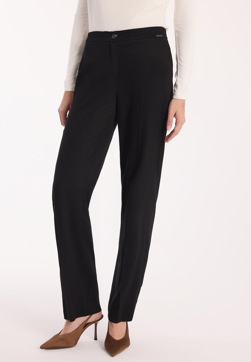 Kobieta ubrana w czarne dopasowane spodnie z zapięciem na guziki, w połączeniu z białą bluzką z długim rękawem oraz brązowymi szpilkami z odkrytym palcem.