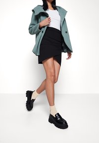 Groene wollen mixjas met een capuchon over een witte top, gecombineerd met een zwarte asymmetrische rok en zwarte chunky instapschoenen met beige sokken.