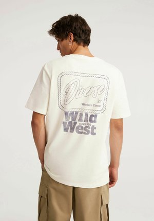 Młody mężczyzna z krótkimi ciemnymi włosami ubrany w kremową koszulkę z napisem "Diverse Wild West estd. 1993" na plecach oraz beżowe krótkie spodnie typu cargo.