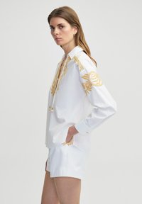 Camicia bianca con ricami dorati, dotata di colletto e lacci. Abbinata a pantaloni corti bianchi, che mostrano una vestibilità ampia e rilassata.