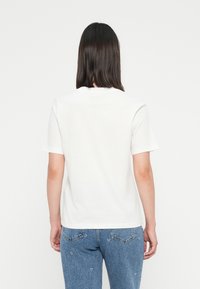 Kvinne med rett, mørkt hår iført en enkel hvit T-skjorte med korte ermer og blå jeans, stående med ryggen til en hvit bakgrunn.