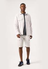 Chemise boutonnée beige clair avec deux poches poitrine, portée sur un t-shirt à rayures foncées. Shorts cargo blancs et baskets bleu foncé avec des rayures.