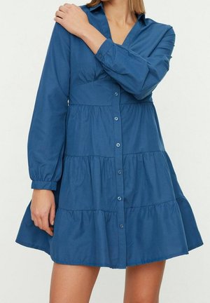 Vrouw in een blauwe, lange mouwen, geknoopte jurk met gelaagde rok, staand met één hand die de tegenovergestelde schouder aanraakt.