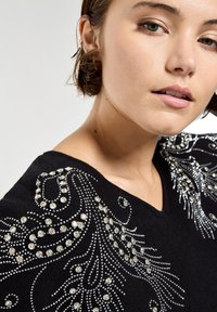 Czarny sweter z misternymi zdobieniami z srebrnych cyrkonii w kwiatowe i spiralne wzory, z dekoltem w kształcie litery V.