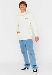 Crème hoodie met een voorzak en een capuchon met trekkoord, gecombineerd met lichtblauwe jeans en zwarte sneakers met witte accenten.