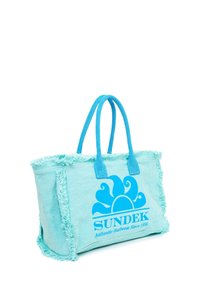 Bolsa de lona azul claro con bordes deshilachados, que presenta un gran logo de sol azul y el texto "SUNDEK Ropa de Surf Auténtica Desde 1958" en el lateral.