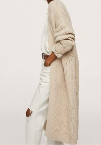 Beżowy dziergany kardigan z fakturowanym wzorem, otwarty z przodu, z długimi rękawami i luźnym krojem. Noszony na białej bluzce i dżinsach.