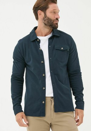 Navyhemden aus sanftem Stoff mit einer Brusttasche, langen Ärmeln und einem Kragen, kombiniert mit einem weißen Unterhemd und beigen Hosen.