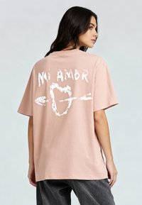 T-shirt en coton rose à coupe décontractée. Le dos arbore un texte graphique blanc "Mi Amor" et un cœur transpercé par une flèche, ajoutant un détail visuel notable.