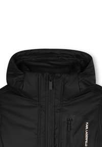 Doudoune noire avec un col montant, fermeture éclair à l'avant et une poche poitrine zippée arborant un logo. Texture lisse et brillante.