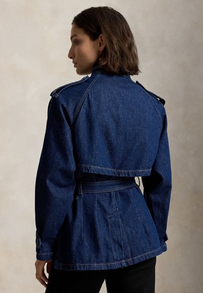 Ženska z ramensko dolgo pričesko, oblečena v temno modro jeans jakno z pasom in epolete, videna od zadaj na enostavni podlagi.
