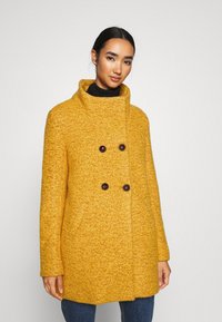 Cappotto in misto lana giallo con alto colletto, chiusura a doppio petto con bottoni e motivo testurizzato. Presenta due tasche laterali.