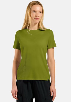 Woman with long blonde hair wearing an olive green short-sleeve shirt and black pants, standing against a plain light background.