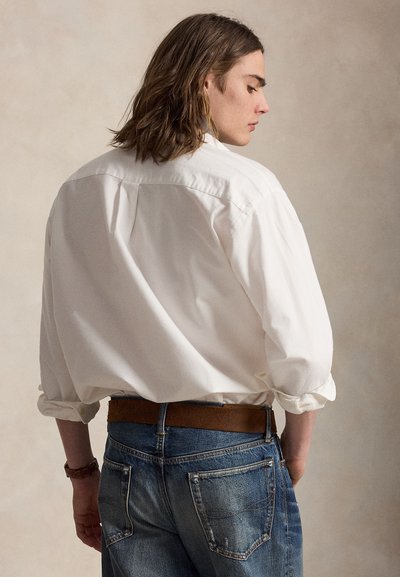 Junge Person mit schulterlangem braunem Haar trägt ein locker sitzendes weißes Hemd und blaue Jeans mit braunem Gürtel, steht seitlich vor einem schlichten Hintergrund.