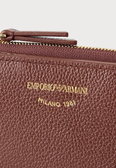 Κοντινή λήψη καφέ υφασμένης δερμάτινης θήκης με χρυσό φερμουάρ και τη λέξη "EMPORIO ARMANI MILANO 1981" ανάγλυφη με χρυσά γράμματα.