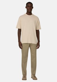 Jeune homme debout portant une chemise beige à manches courtes surdimensionnée, un pantalon marron clair et des baskets en daim assorties, sur un fond uni.