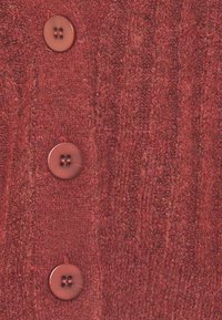 Röd stickad cardigan med tre runda, släta knappar. Texturen är mjuk med ett synligt ribbat mönster och små inslag av mörkare röd.