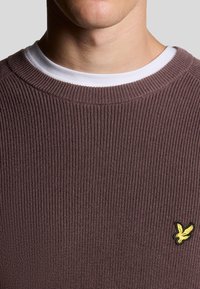 Bruine geribbelde gebreide sweater met een ronde hals en een witte binnenkraag. Gele geborduurde vogel logo op de linkerkant van de borst. Zachte textuur.