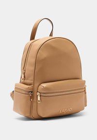 Mochila de cuero color canela con forma redondeada, dos bolsillos delanteros con cremallera, detalles de herrajes dorados y logotipo en relieve en la parte inferior.