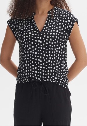 Femme portant un haut noir sans manches avec des motifs carrés blancs et un pantalon noir, debout les mains dans le dos.