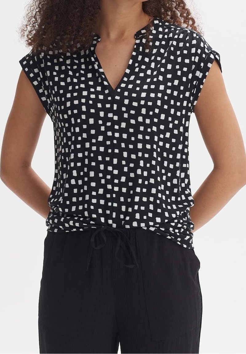 Femme portant un haut noir sans manches avec des motifs carrés blancs et un pantalon noir, debout les mains dans le dos.