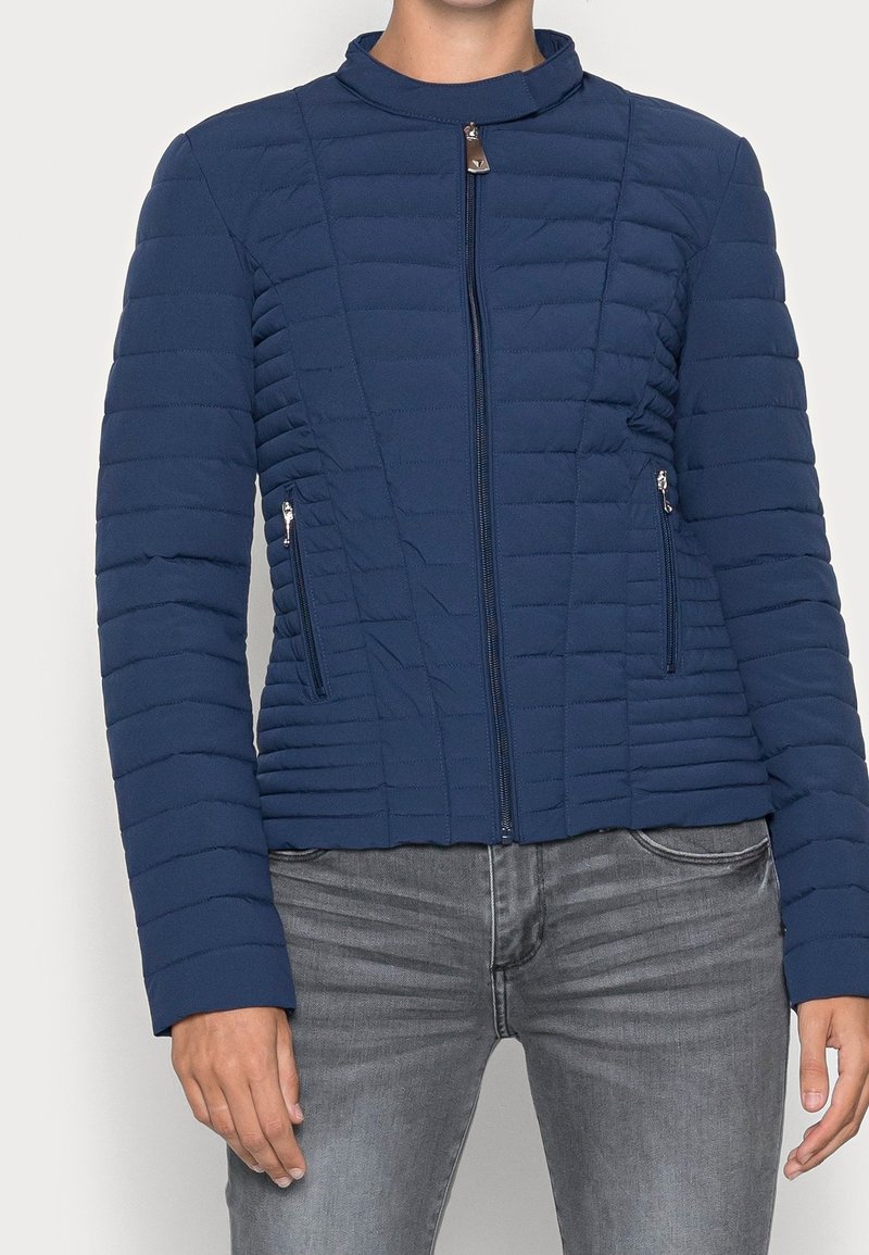 Giacca trapuntata blu con colletto alto, dotata di una chiusura frontale con zip e due tasche laterali. Il materiale ha una texture liscia con cuciture orizzontali.