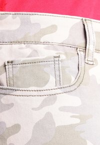 Kamouflagemönstrad tyg i ljusa beige toner. Har en bakficka med sömnadsdetaljer, fäst vid en ljusröd topp.