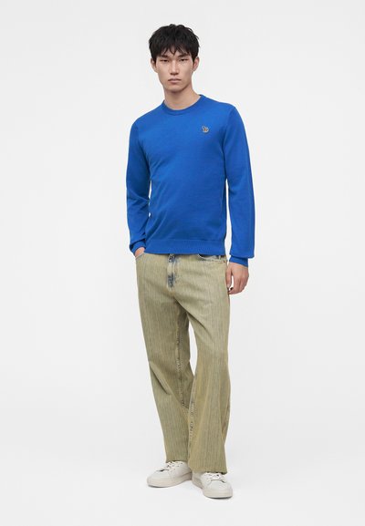 Jeune homme portant une chemise bleue à manches longues, un pantalon beige à jambes larges et des baskets blanches, debout sur un fond blanc uni.