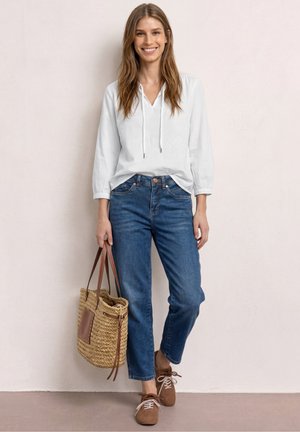 Hymyilevä nainen valkoisessa paidassa, sinisissä farkuissa ja ruskeissa kengissä seisoo ja pitää käsissään punottua olkikassia vaalean seinän edessä.