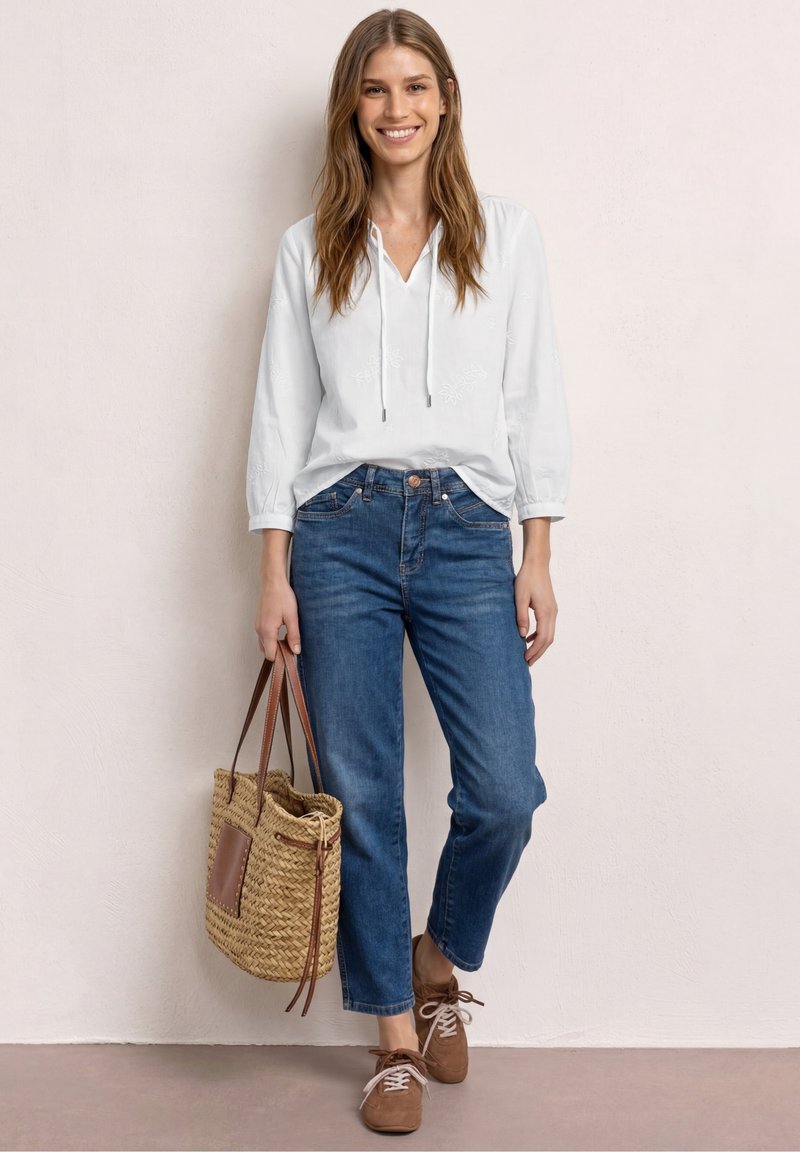 Femme souriante en blouse blanche, jean bleu et chaussures marron, debout tenant un sac cabas en paille tressée contre un mur pâle.