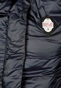 Marinblå puffjacka med quiltad textur, med en filtlogga på svart bakgrund. Slätt, glänsande tyg med en tät design.