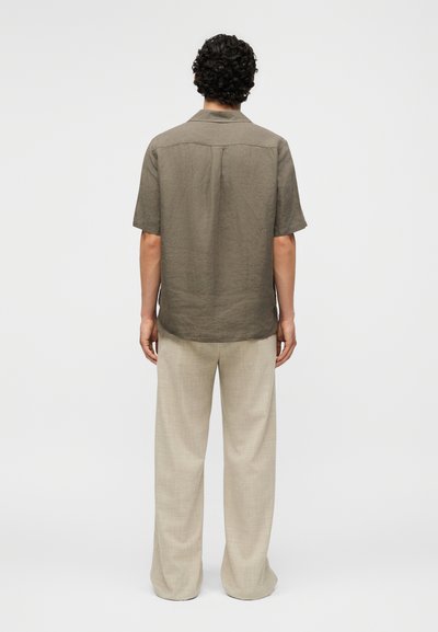 Άνδρας με σγουρά μαλλιά που φοράει άνετο ελιάς κοντομάνικο πουκάμισο και μπεζ παντελόνια φαρδιάς ποδός, στέκεται με την πλάτη προς τα εμπρός σε απλό φόντο.