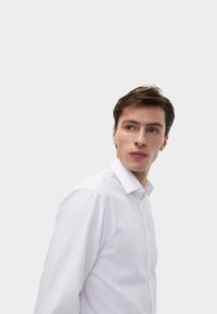 Bílé košilové tričko s pevným límcem, dlouhými rukávy a hladkou texturou, které má na sobě model dívající se do strany na neutrálním pozadí.