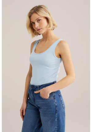 Heledate juustega blond naine, kes kannab helerohelist tankitoppi ja kõrge vöökohaga siniseid pükse, käsi taskus, tavalise tausta ees.