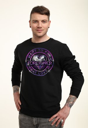 Sort sweatshirt med et rundt grafik, der har et hjerte-design, lilla accenter og teksten "I WANT TO KNOW WHAT LOVE IS FOREIGNER."