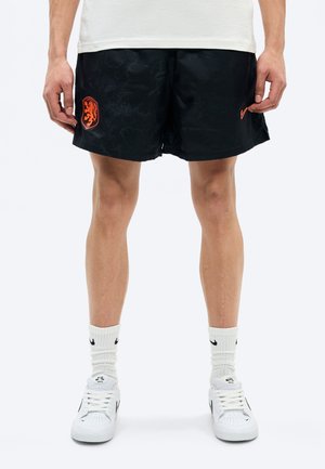 Mężczyzna w białych butach Nike, białych skarpetkach Nike, czarnych szortach z pomarańczowym herbem lwa i logo Swoosh, stojący na białym tle.