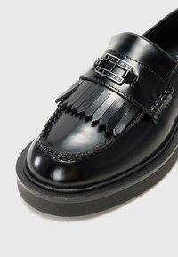 Mocassino in pelle nera con design frangiato, arricchito da una placca logo in argento e cuciture decorative. Suola spessa in gomma nera.
