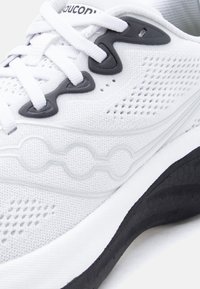 Λευκό αθλητικό παπούτσι Saucony με μαύρες λεπτομέρειες, ανώτερο μέρος από διαπνέον δίχτυ και επενδεδυμένη σόλα, εμφανιζόμενο σε κοντινή λεπτομέρεια.