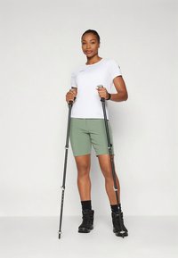 Balta mitrumu atgrūdoša T-krekla krekla un zaļi šorti. Melni pārgājienu zābaki ar teksturētu zolīšu virsmu. Tur divas pārgājienu nūjas ar gumijas rokturiem.