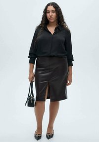 Sort silke blouse og knælang brun lædernederdel med en frontslids, parret med en lille sort håndtaske og spidse sorte sko.