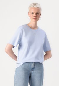 Light blue short-sleeve t-shirt made of soft fabric, featuring a round neckline and a small embroidered logo on the chest. Paired with blue jeans.