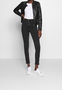 Zwarte leren bomberjack met een ronde kraag, gecombineerd met hooggehupte zwarte skinny jeans met knoopsluiting en rafelige zoom. Witte sneakers.