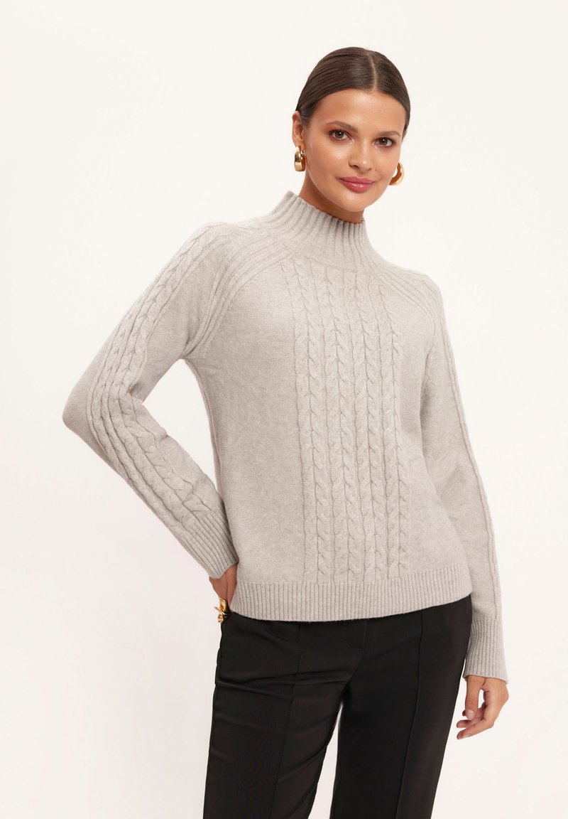 Jasnoszary sweter z prążkowanym wysokim kołnierzem oraz skomplikowanym wzorem w warkocze biegnącym w dół środka i na rękawach, połączony z czarnymi spodniami.