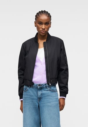 Donna con capelli intrecciati che indossa una giacca bomber nera, un cardigan lilla, una maglietta bianca e jeans a vita alta azzurri davanti a uno sfondo semplice.